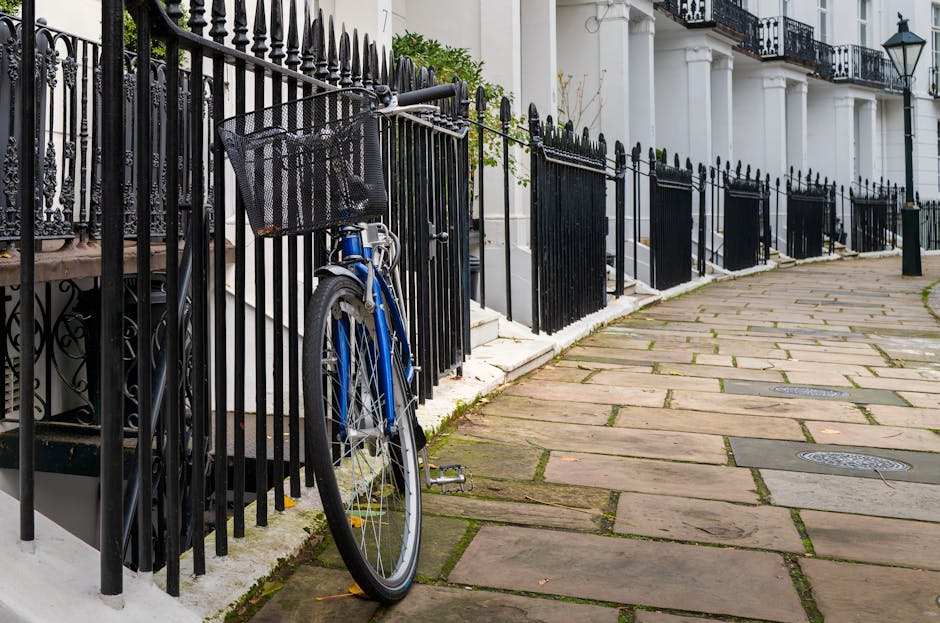 A blue bicycle with a black front basket is parked against a black metal fence along a paved sidewalk in a residential area. The fence features vertical bars and ornate finials, enclosing white terrac