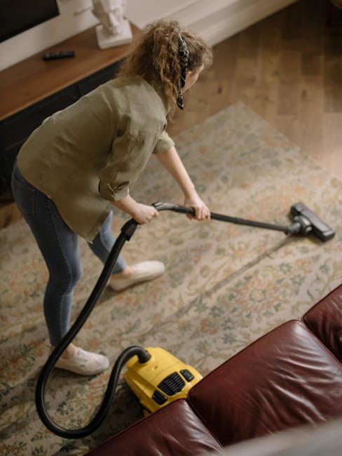 A professional cleaner wearing a white protective suit, gloves, and shoe covers is performing deep cleaning on a beige carpet in a bright, well-lit living room. The cleaner is using a large vacuum hos